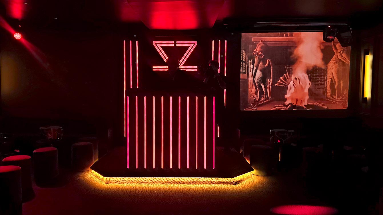 Salle de l'étage du Zouzou Pigalle avec cabine DJ néon rouge, poufs rouges et écran projetant des créatures fantastiques.