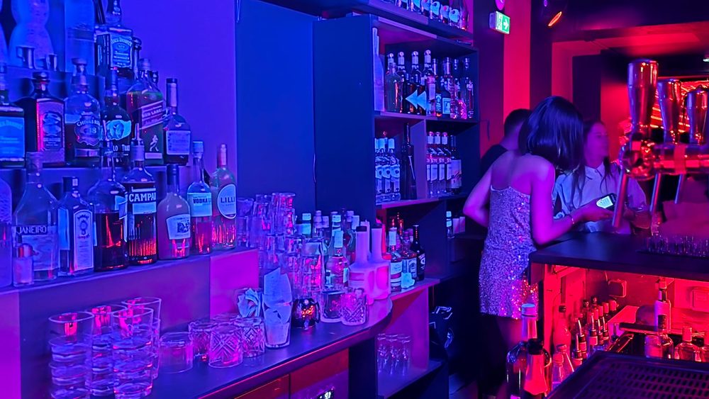 Bar du Zouzou Pigalle éclairé en rose et violet, avec étagères de bouteilles, verres et tireuses à bière, et une serveuse en robe à sequins.