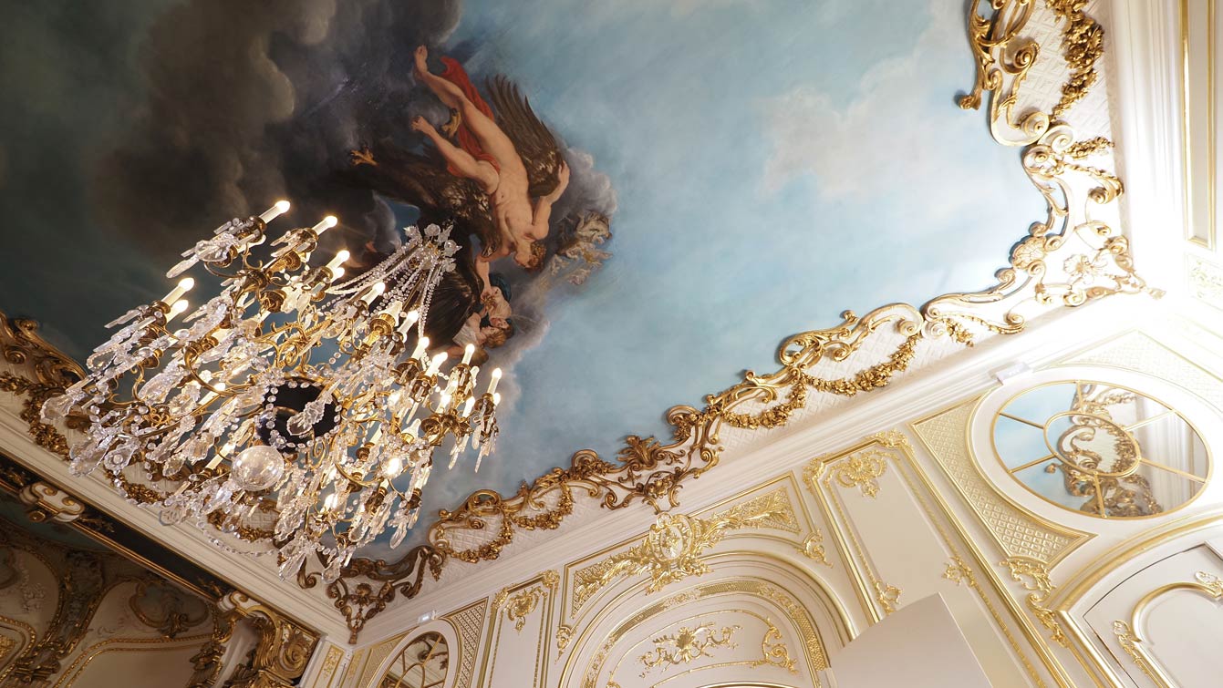 Plafond peint d'un ciel bleu avec un personnage mythologique, encadré de dorures et éclairé par un lustre en cristal, à l'Hôtel Le Marois