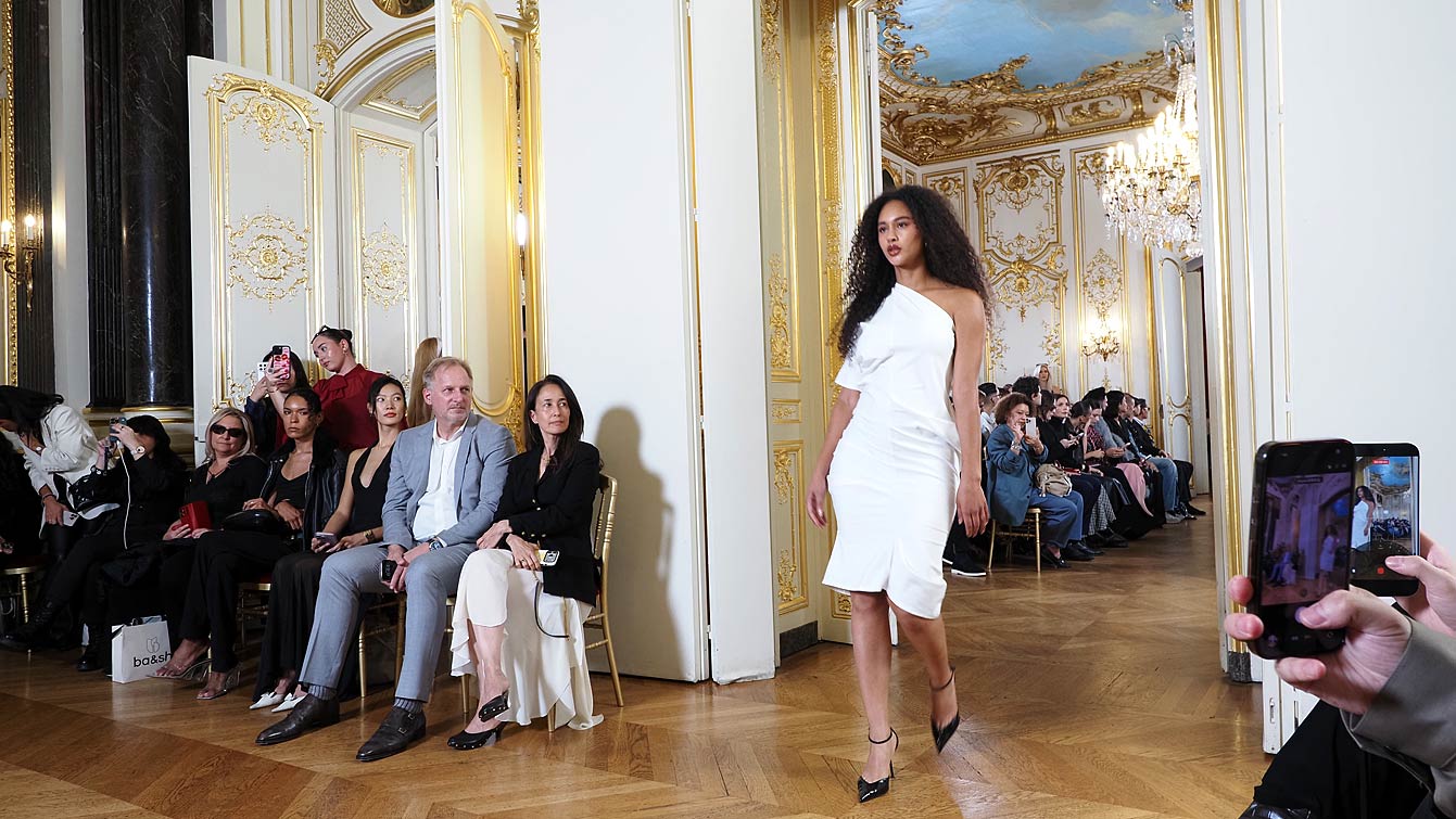 Mannequin en robe blanche asymétrique défilant entre les invités assis dans les salons dorés de l'Hôtel Le Marois, pendant le défilé NEXT ASIA