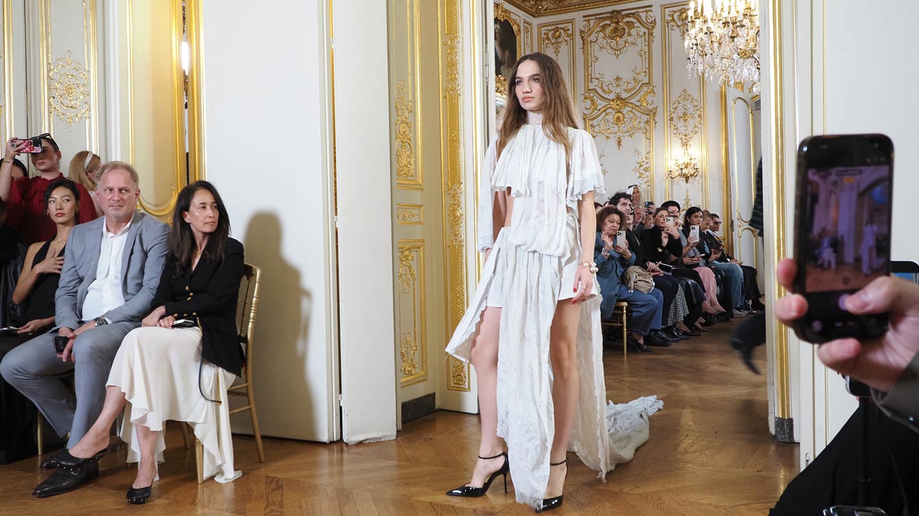 Mannequin en longue robe blanche à volants défilant sur le parquet d'un salon de l'Hôtel Le Marois, devant le premier rang, lors du défilé NEXT ASIA