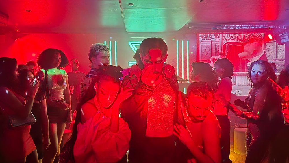 Groupe de danseurs en résille avançant au milieu des spectateurs sous lumière rouge au Zouzou Pigalle. Peu à peu, les numéros s'espacent et la nuit bascule vers le club.