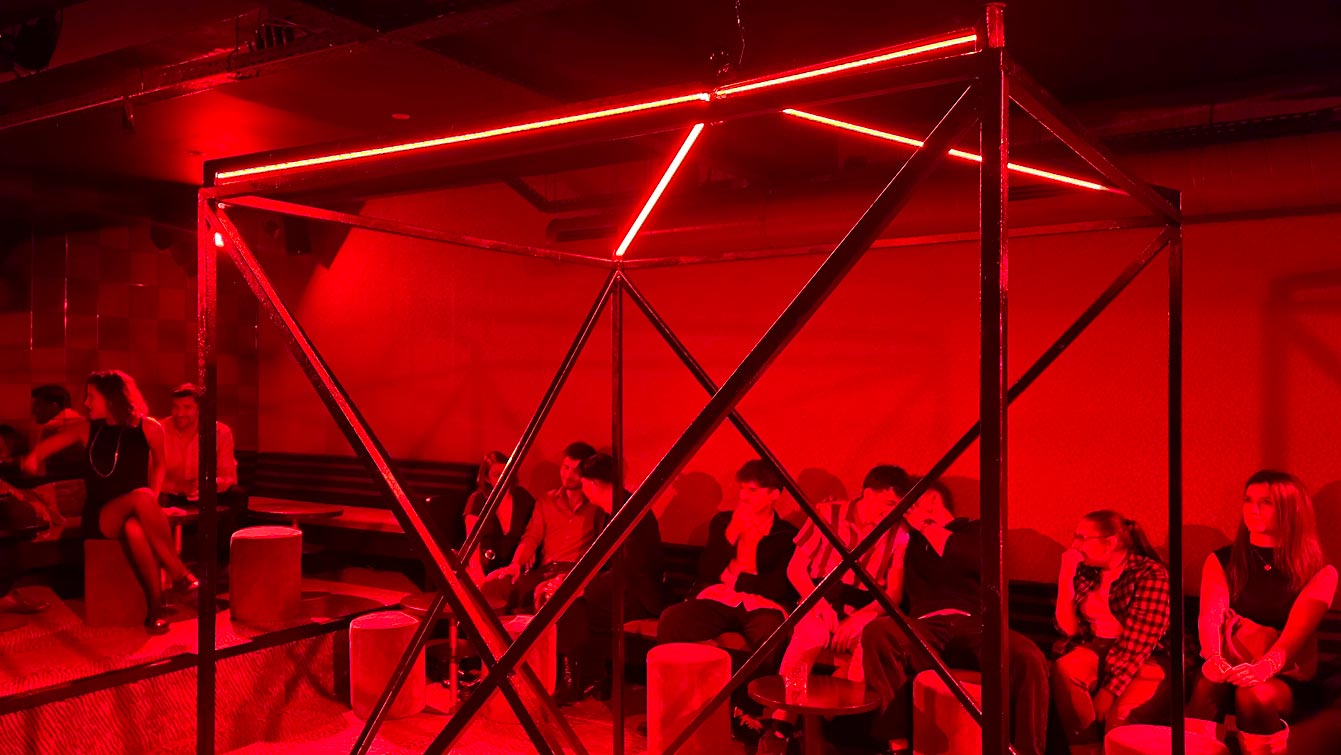 Cage métallique éclairée de néons rouges dans la salle du sous-sol du Zouzou Pigalle, public assis autour.