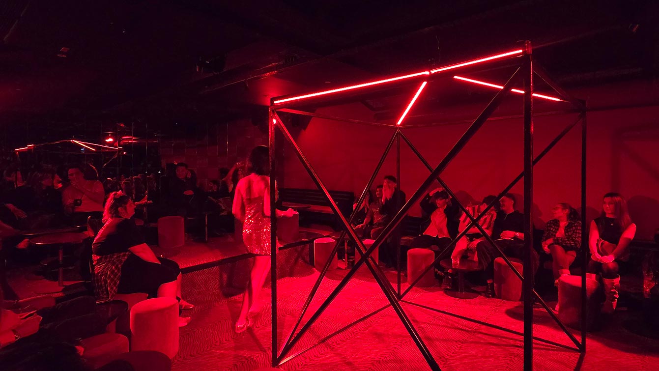 Cage métallique éclairée de néons rouges dans la salle du sous-sol du Zouzou Pigalle, public assis autour.