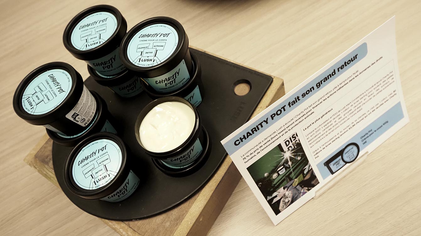 Pots de crème Charity Pot de Lush à couvercle noir et étiquette bleue, dont un ouvert sur une crème blanche, sur un présentoir en bois